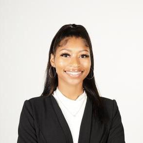 Demi Ward smiles at the camera in front of a white wall, she wears silver hoop earrings and a black blazer with a white shirt underneath.