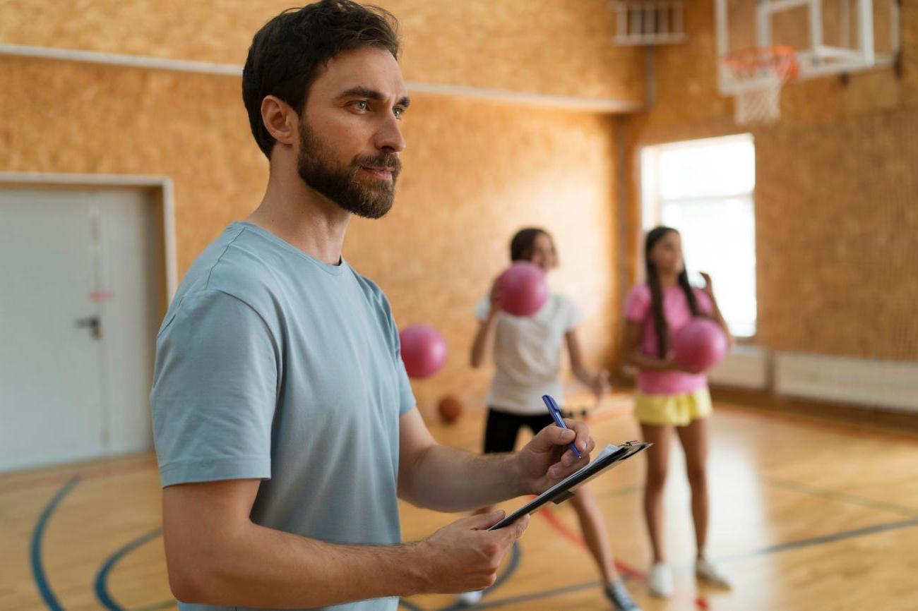 Man writing on clipboard with children in background throwing dodgeballs. Image by Freepik.